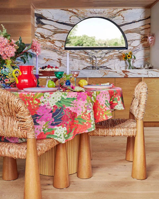 Bush blossom round tablecloth with chunky timber chairs sat around table, styled with red jug, colourful glassware, plates, cake stand and blossom flowers in vase. Marble counter top and wall in background with arch window. 