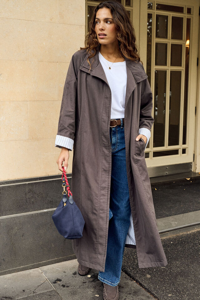 A woman stands on a city sidewalk wearing a long taupe-brown trench coat over a white top and blue jeans, holding a navy blue handbag.