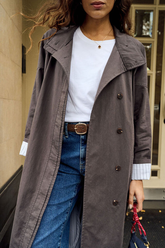A woman wearing a long, double-breasted taupe-brown trench coat over a white crewneck t-shirt, blue jeans, and a brown belt, holding a small red and black handbag.