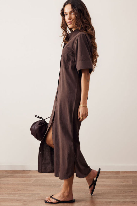 A woman modeling a dark brown short-sleeve midi shirt dress with a high collar, shown in profile against a neutral off-white wall and light wood floor.
