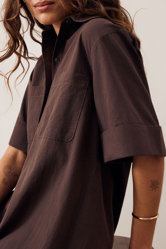 A close-up of a model wearing a dark brown short-sleeve button-up shirt with rolled cuffs and a chest pocket, set against a plain light background.