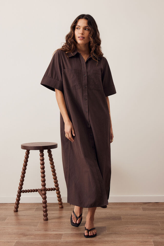 A woman modeling a dark brown short-sleeve button-front midi dress with a relaxed, oversized fit, standing in a room with a wooden floor and a wooden stool.