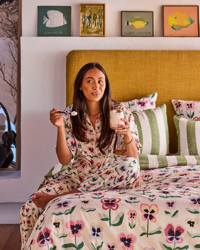 A woman sits on a bed wearing a matching floral pajama set, holding a spoon and a glass of milk in a bedroom with a mustard yellow headboard and floral bedding.