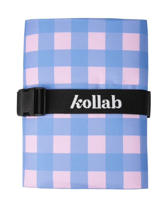 A folded picnic mat in a black strap with the brand name 'kollab' in white, featuring a light blue and pink gingham check pattern, on a plain white background.