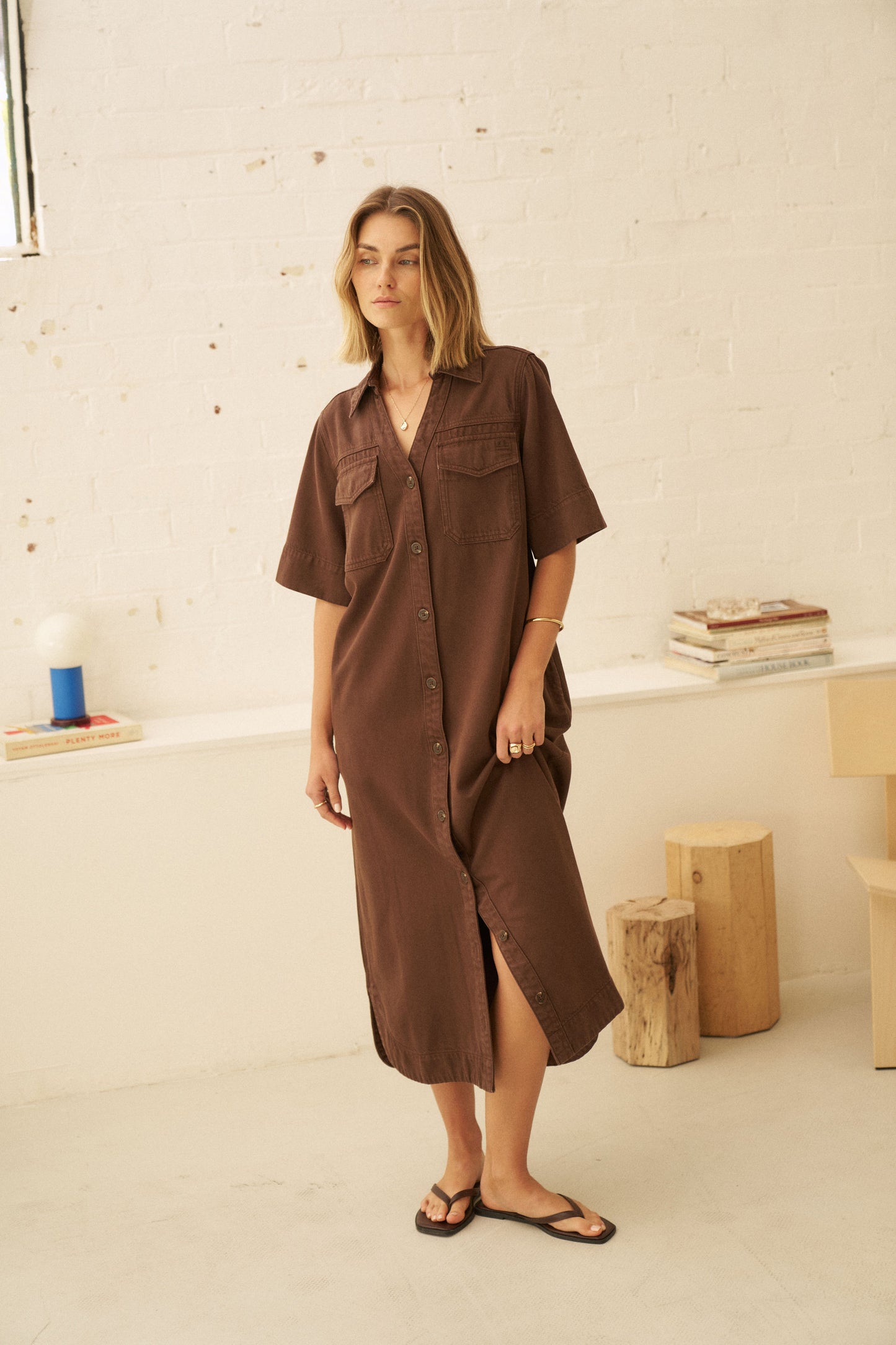 A woman wearing a brown short-sleeve button-up shirt dress with chest pockets, standing in a bright, minimalist room with a white brick wall background.