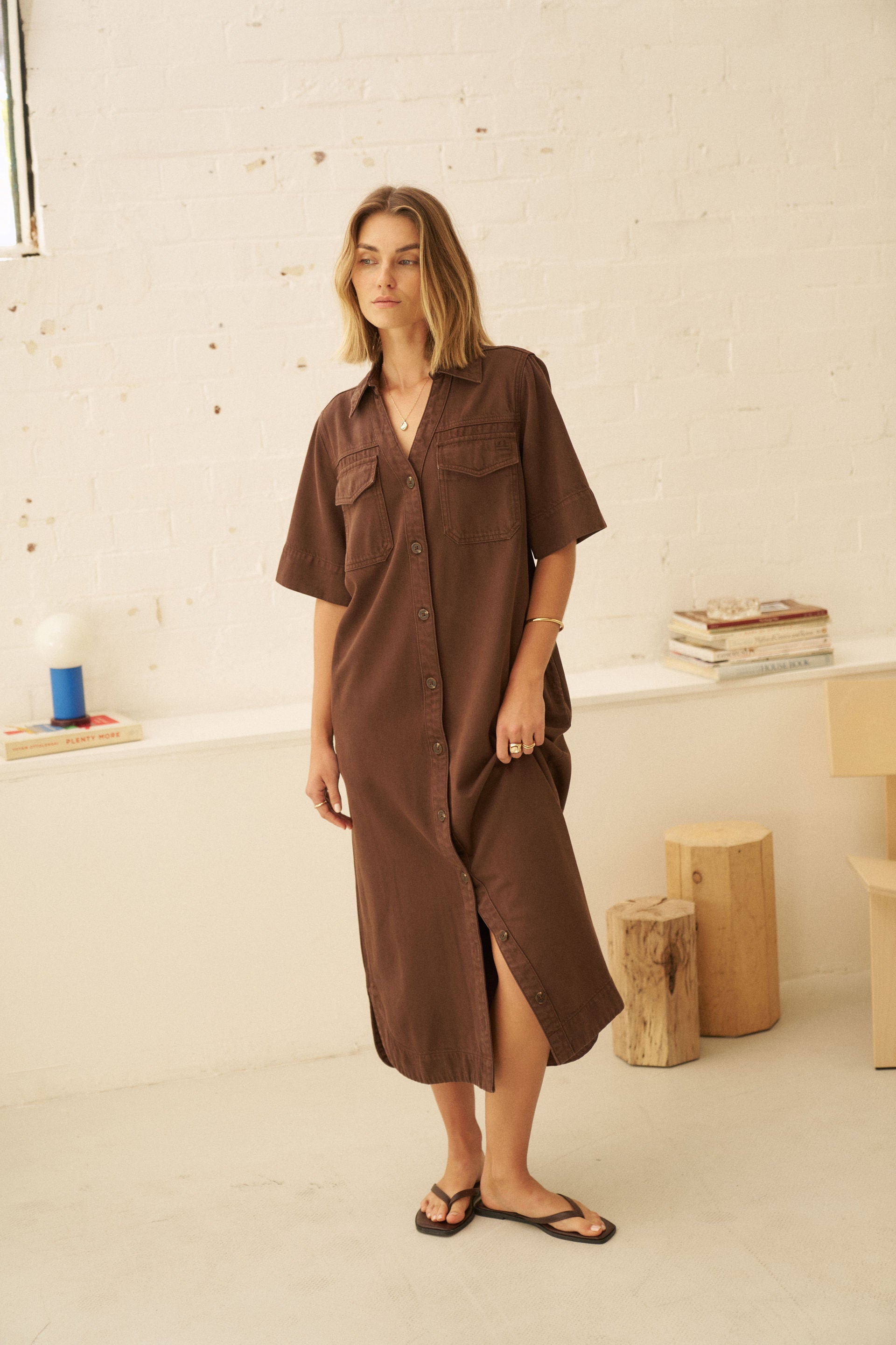 A woman wearing a brown short-sleeve button-up shirt dress with chest pockets, standing in a bright, minimalist room with a white brick wall background.
