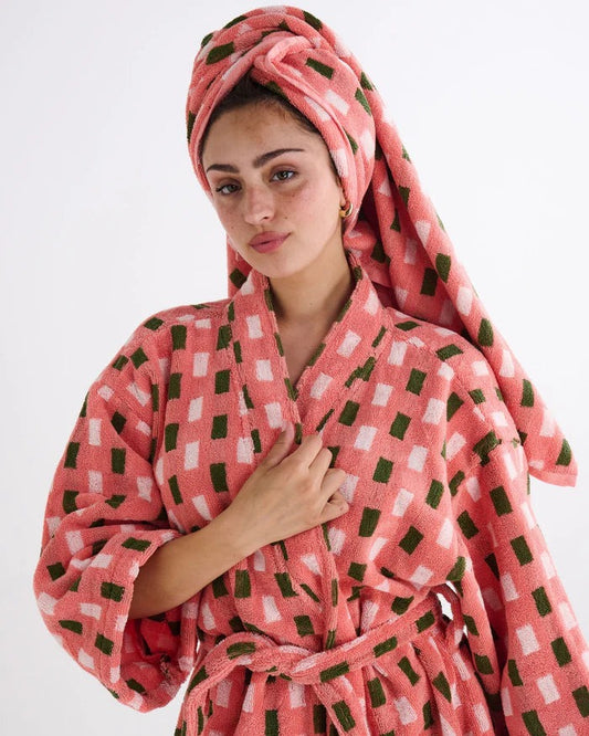 A woman wearing a pink bathrobe with a green and white checkered pattern, with a matching pink towel wrapped around her head, against a plain white background.