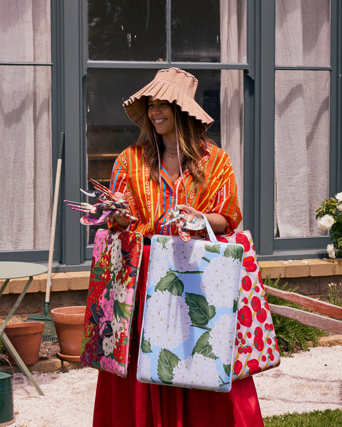 A woman wearing a wide-brimmed straw hat, an orange patterned top, and a red skirt stands outdoors in front of a house window, holding three colorful strawberry-themed tote bags.
