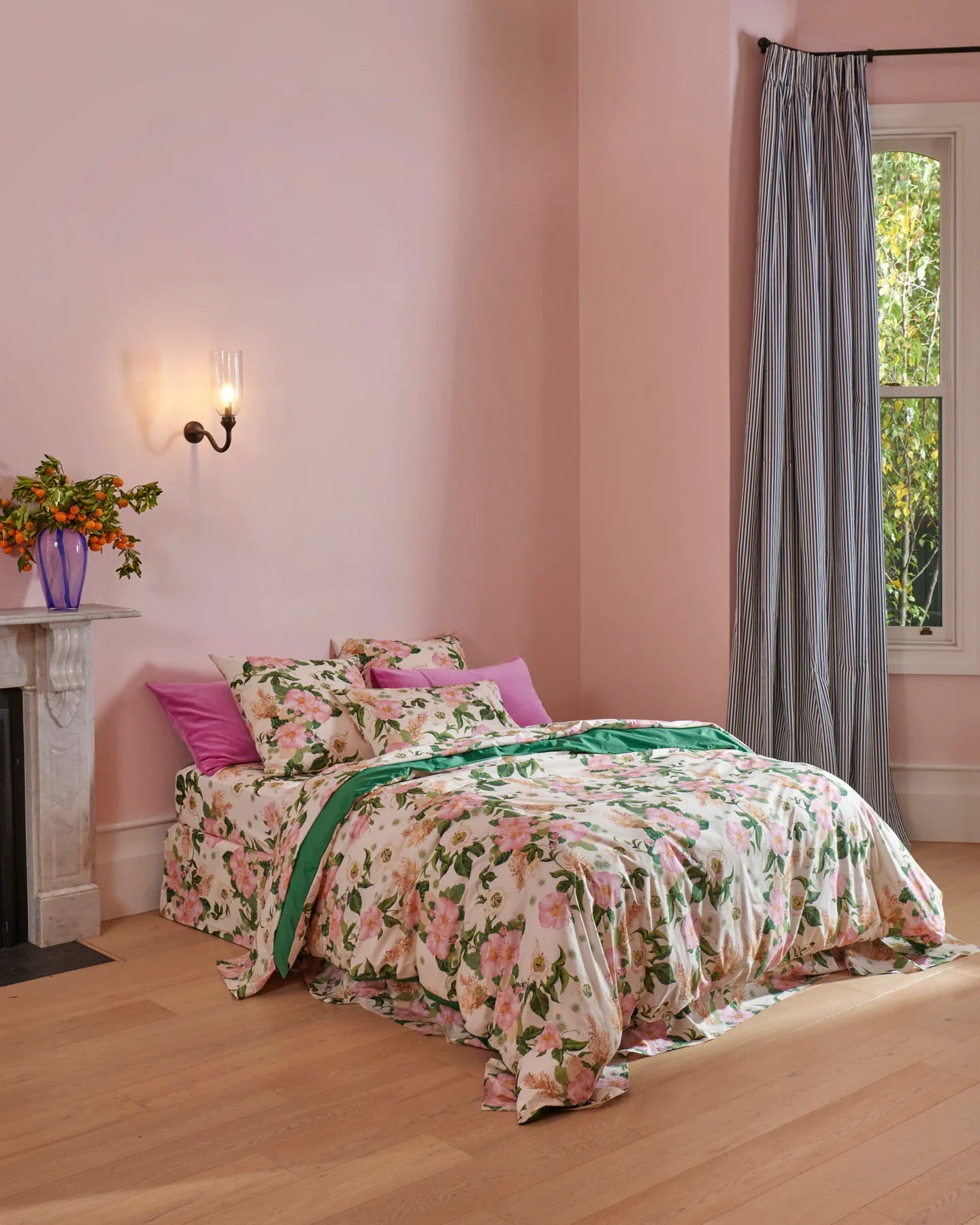 Bed styled with cream base quilt cover and pink green rose pattern, quilt cover has solid green backing. Pillows are matching print and also pink velvet standard pillowcases. Marble fire place sat next to bed with glass vase and orange greenery filling vase. 
