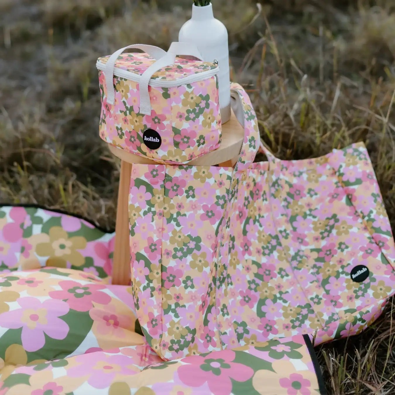 A coordinated picnic setup outdoors in grass, featuring a pink floral blanket, a matching tote bag, a small floral lunch bag, and a white bottle on a wooden stand.