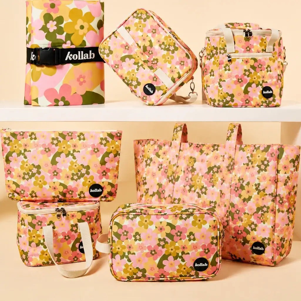 A coordinated set of floral fabric bags and pouches displayed on white shelves against a warm beige background. The set includes a rolled sleeve, a small pouch, a backpack-style bag, a tote bag, a zippered pouch, and a larger tote, all in a pink, yellow, green, and white floral print.