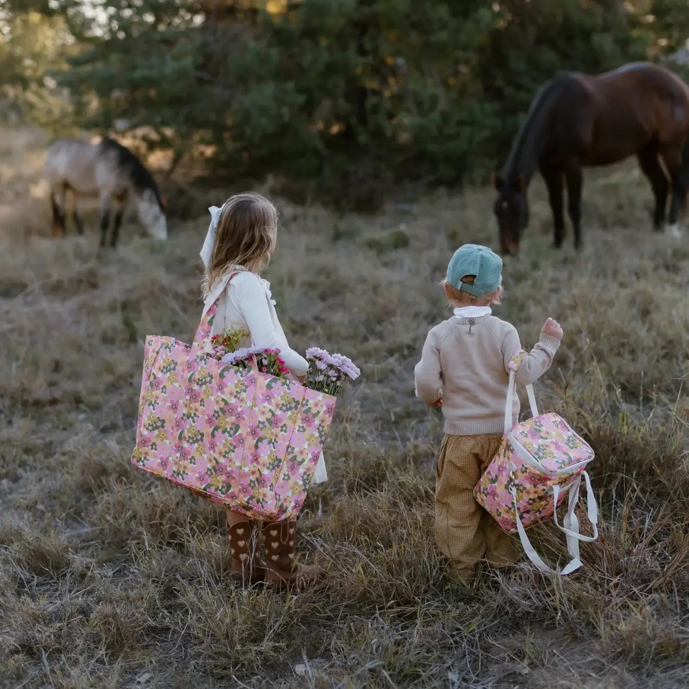 Two children standing in a grassy field with horses in the background, each carrying a pink floral insulated tote bag. The scene is shot from behind in warm natural light.