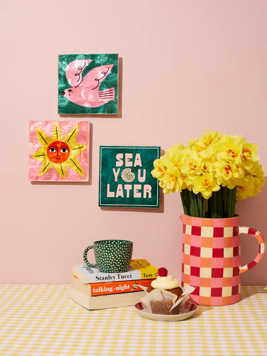 Decorative items including a mug, books, and a vase with flowers on a table against a pink wall with framed pictures.
