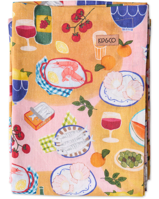 A folded linen tablecloth with a printed design featuring food items like seafood, citrus, melon, olives, and wine glasses, along with a brand logo 'KIP & CO'.