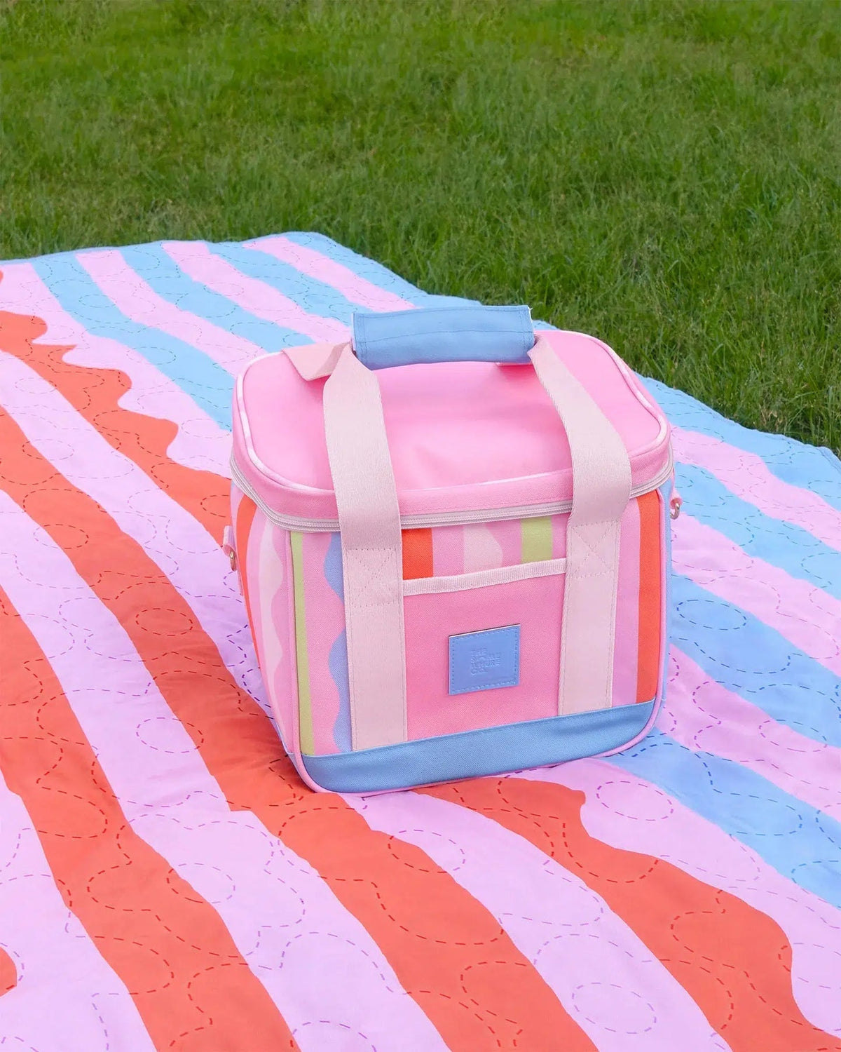A pastel pink cooler bag with light blue trim and cream straps sits on a striped picnic blanket outdoors with green grass in the background.