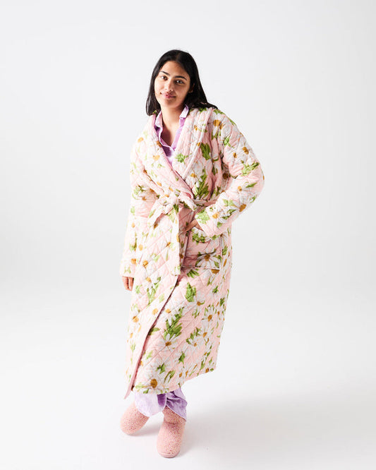 A woman modeling a light pink quilted bathrobe with a green and yellow floral pattern, styled over lavender pajama pants and pink slippers against a white studio background.
