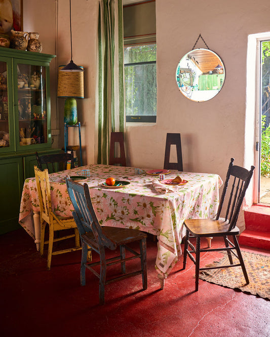A rustic dining room scene featuring a floral tablecloth on a wooden dining table surrounded by vintage wooden chairs, set in a room with a red floor, green curtains, and a round mirror on the wall.