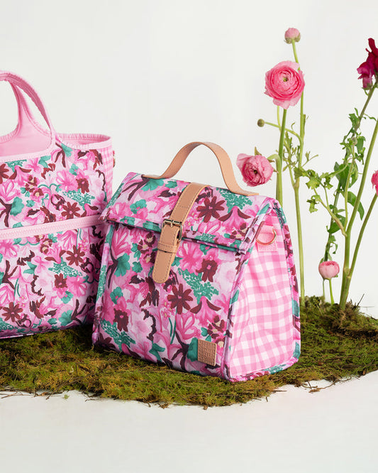 A pink floral and gingham patterned tote bag and matching lunch bag styled on moss with pink flowers against a white background.