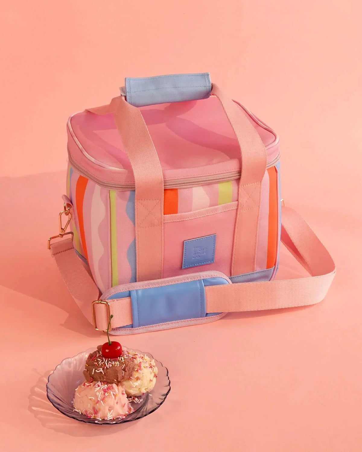 A pastel pink and multicolor insulated lunch bag with a top handle and shoulder strap, styled on a peach-pink background next to a small plate of ice cream with a cherry.
