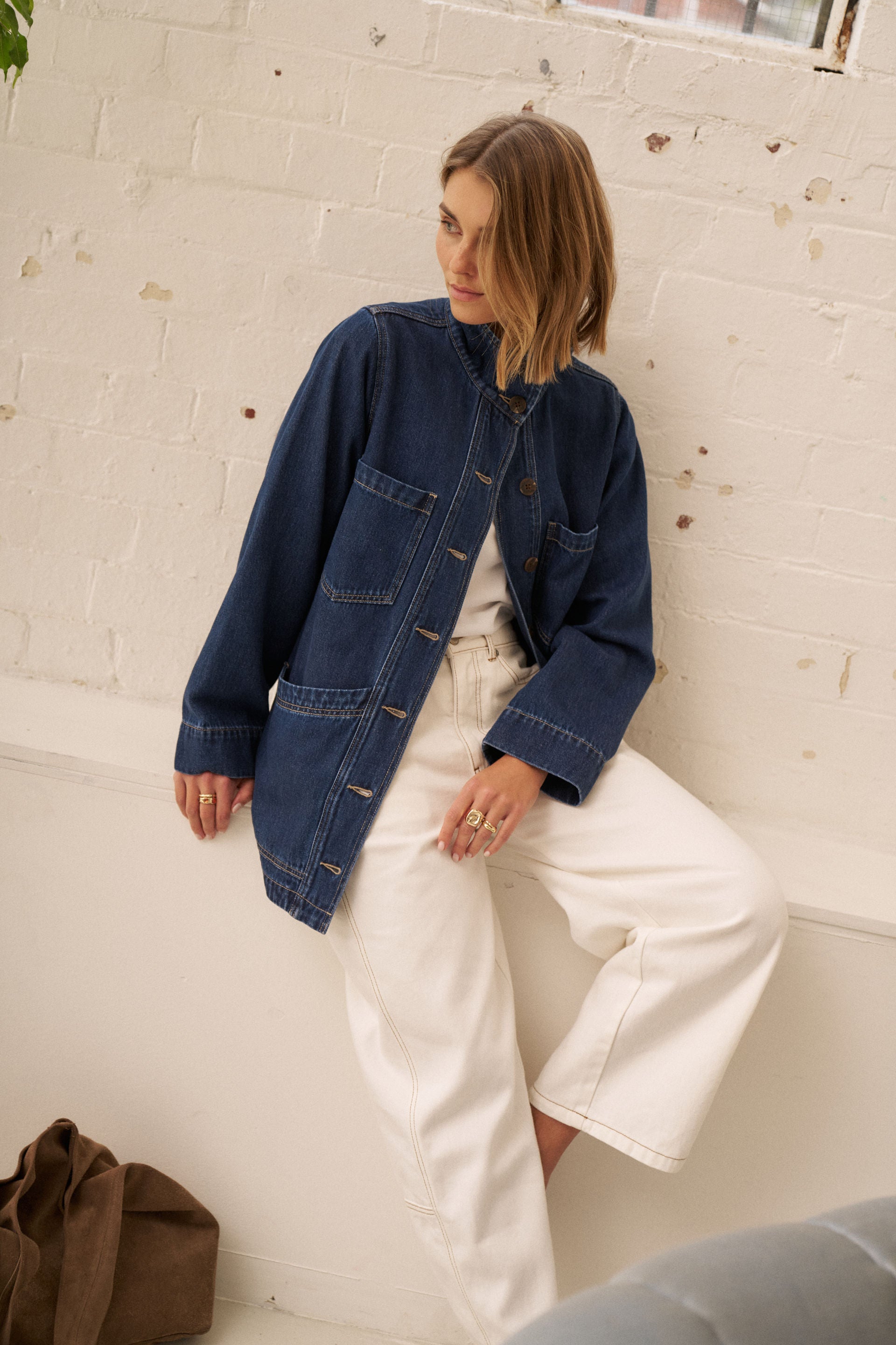 A woman wearing a dark blue denim jacket with toggle closures and white wide-leg pants, leaning against a white brick wall.