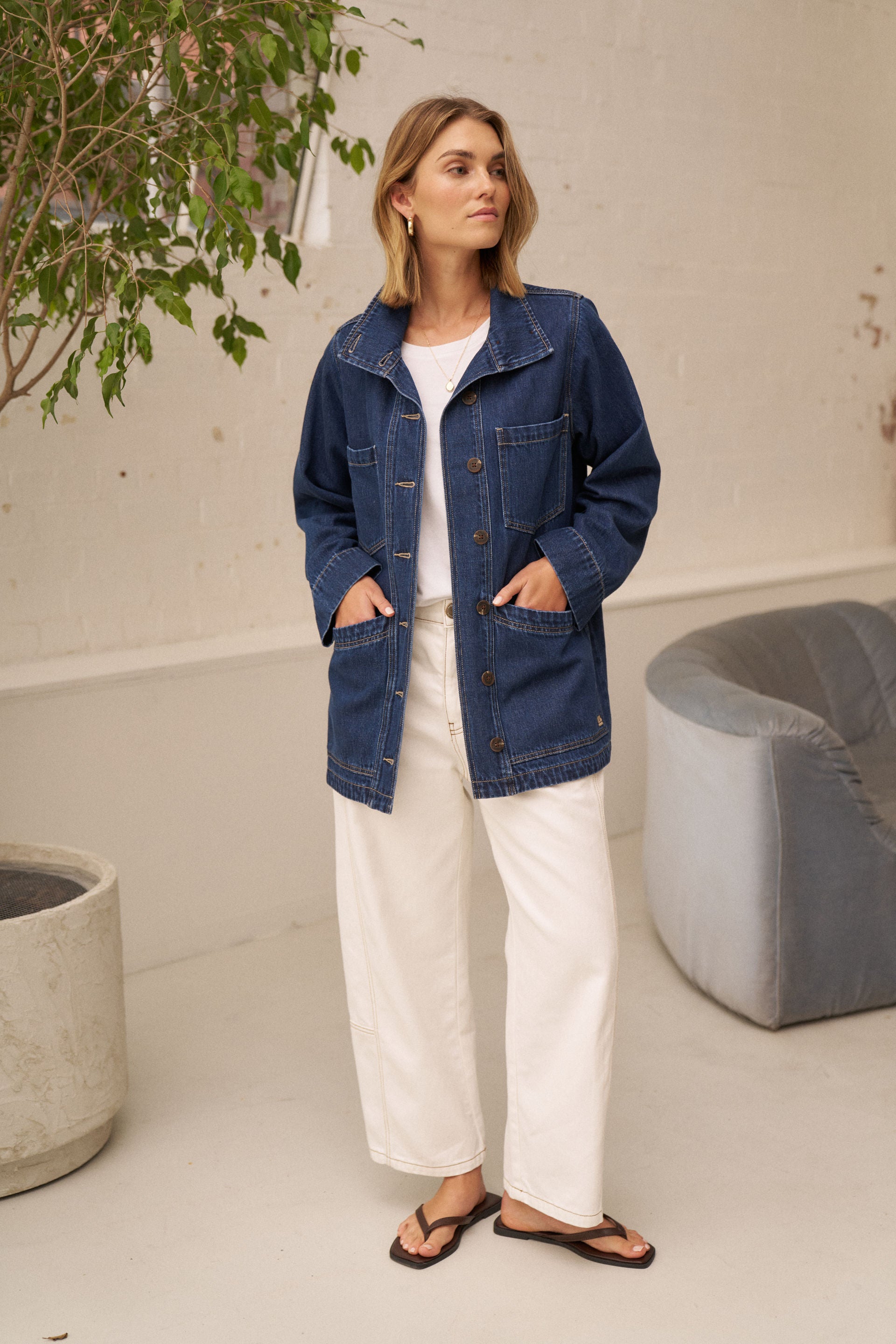 A woman wearing a blue denim utility jacket over a white top and white wide-leg pants, standing in a bright indoor setting with a plant, concrete planter, and gray chair visible.