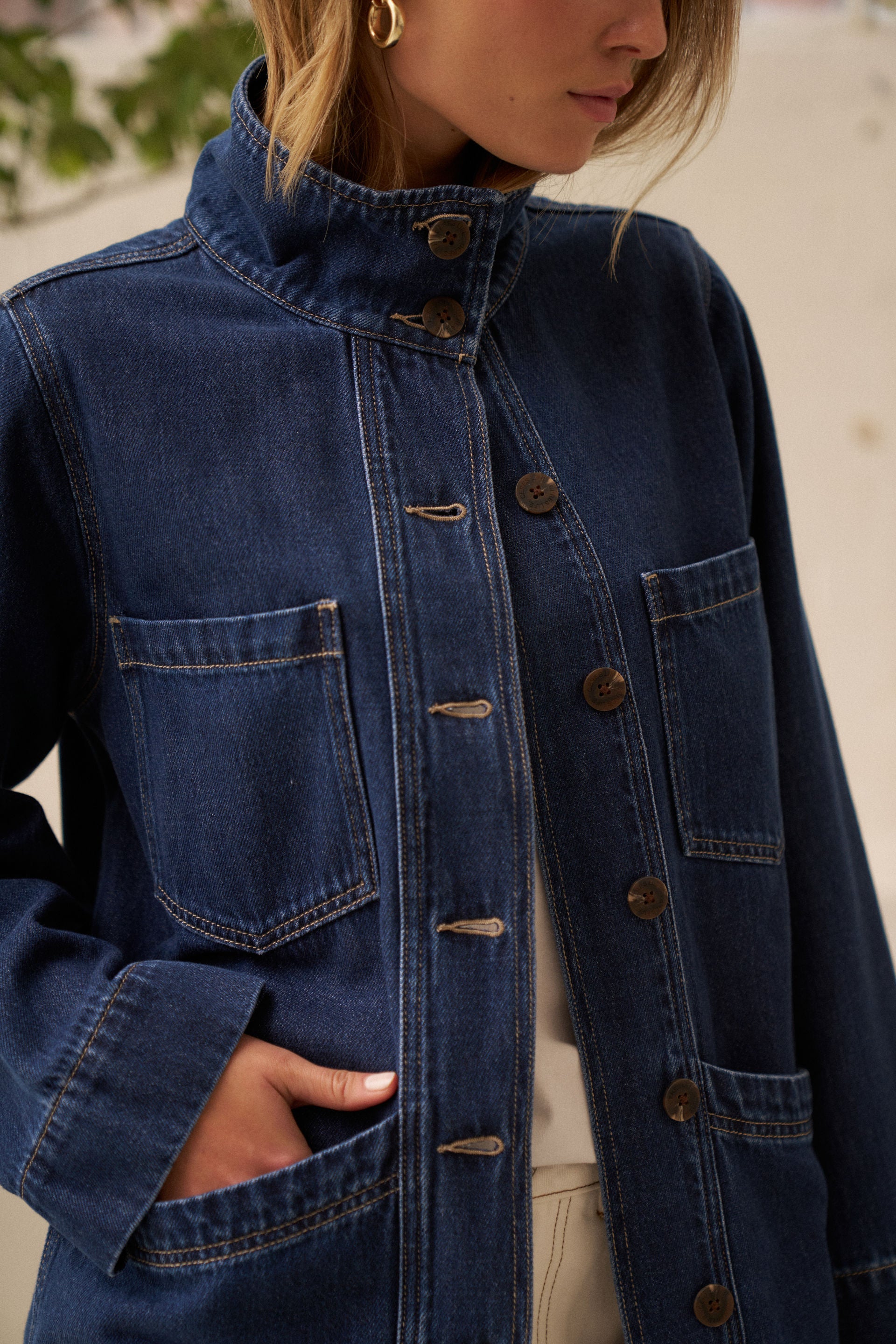 Woman with hand in pocket of denim jacket with two buttons done up at neck and rest left open.
