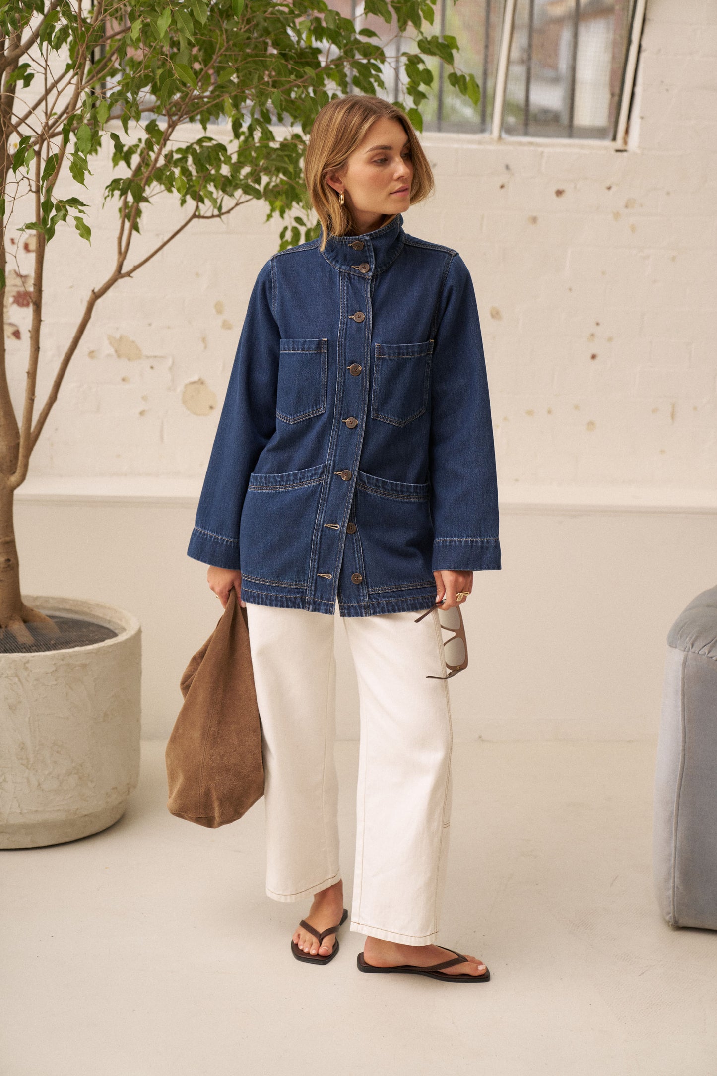 A woman stands indoors wearing a blue denim jacket with a high collar and front buttons, paired with white wide-leg pants and black sandals. She holds a brown tote bag and sunglasses, with a potted tree and a light-colored wall in the background.