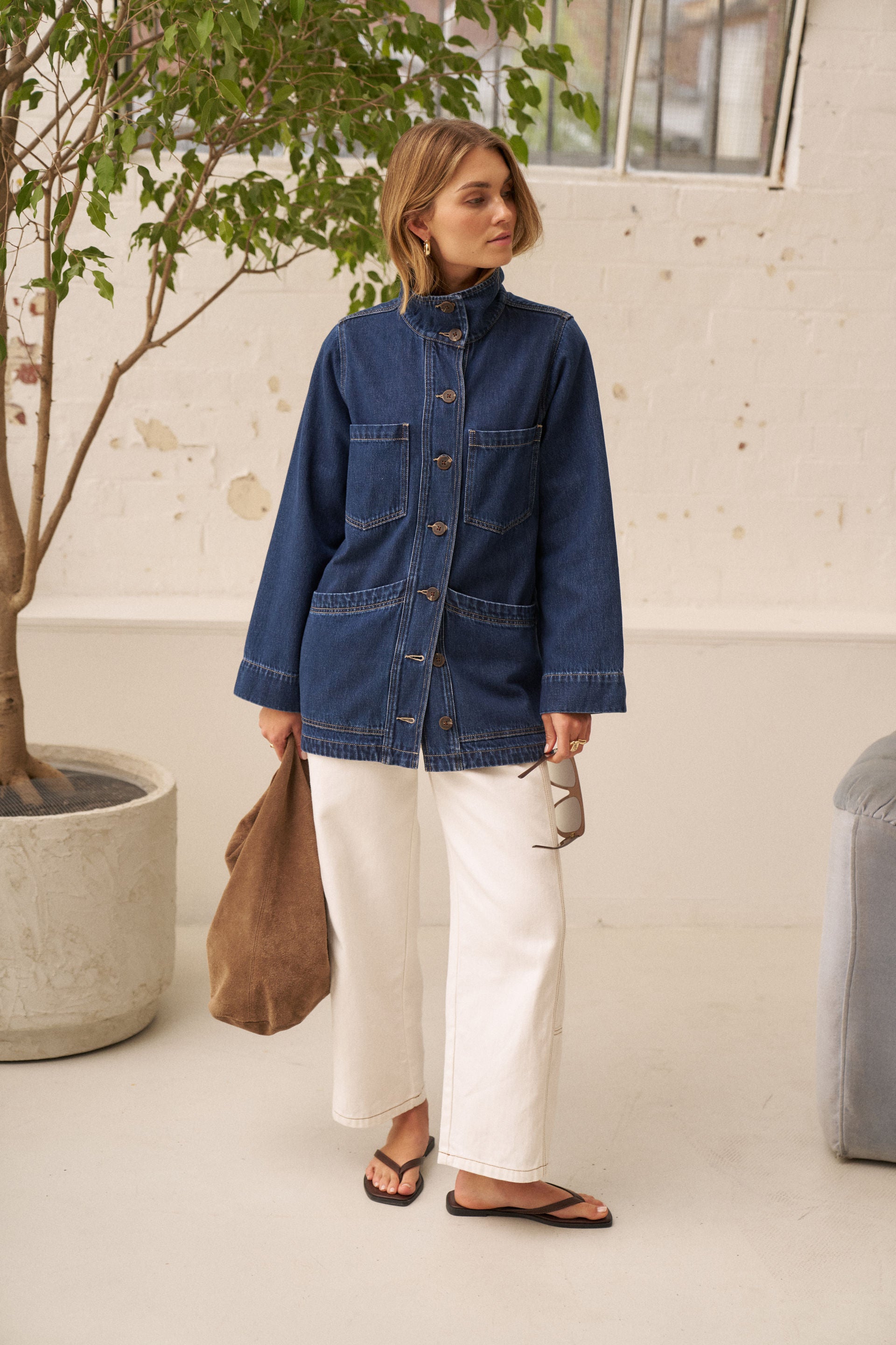 A woman stands indoors wearing a blue denim jacket with a high collar and front buttons, paired with white wide-leg pants and black sandals. She holds a brown tote bag and sunglasses, with a potted tree and a light-colored wall in the background.