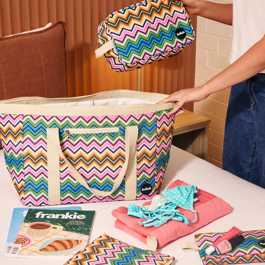 Woman with cosmetic case holding onto tote bag. Magazines, lip glosses sit in front on table. 