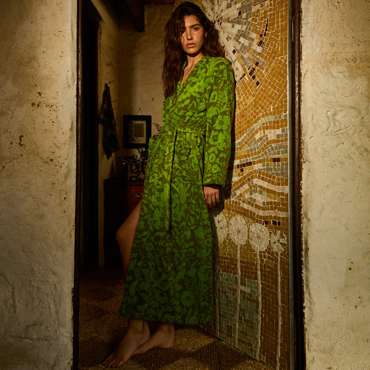 A woman stands barefoot in a rustic doorway wearing a long-sleeved green robe with an abstract leaf-like pattern. The setting features a textured plaster wall and a mosaic tile panel behind her.