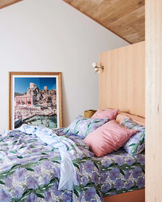 Tumbling flowers printed cotton quilt cover, matching fitted sheet and pillowcases - pattern is white base with blue flowers and green leaf detail. Pink euro pillowcases on bed. Leaning against wall next to bed is image of italian beech and hills. Timber roof, white wall, timber wall with pendant sconce light.