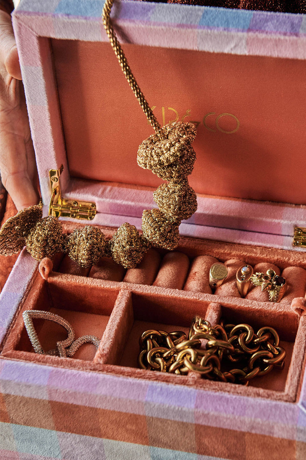 Velvet jewellery box in gingham pattern in assorted pink, blue and orange colours. Gold necklace hangs from lid of box, assorted rings sit upright in ring holder and bracelets cascade out of sections. 