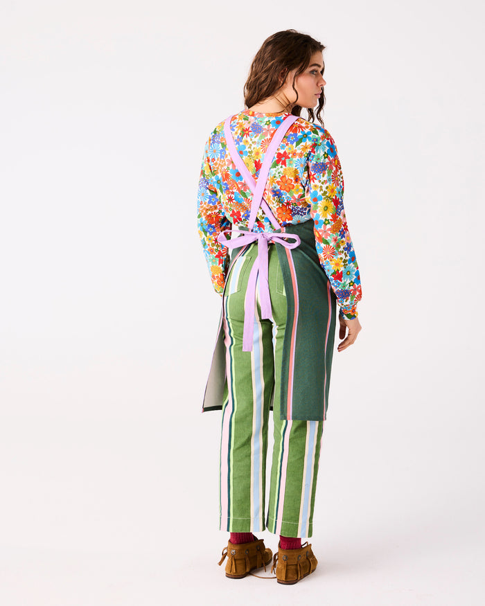 A model is shown from the back wearing a colorful floral long-sleeve top, green striped pants with pink and white side stripes, and a dark green apron with a pink cross-back strap and purple tie, set against a plain white studio background.