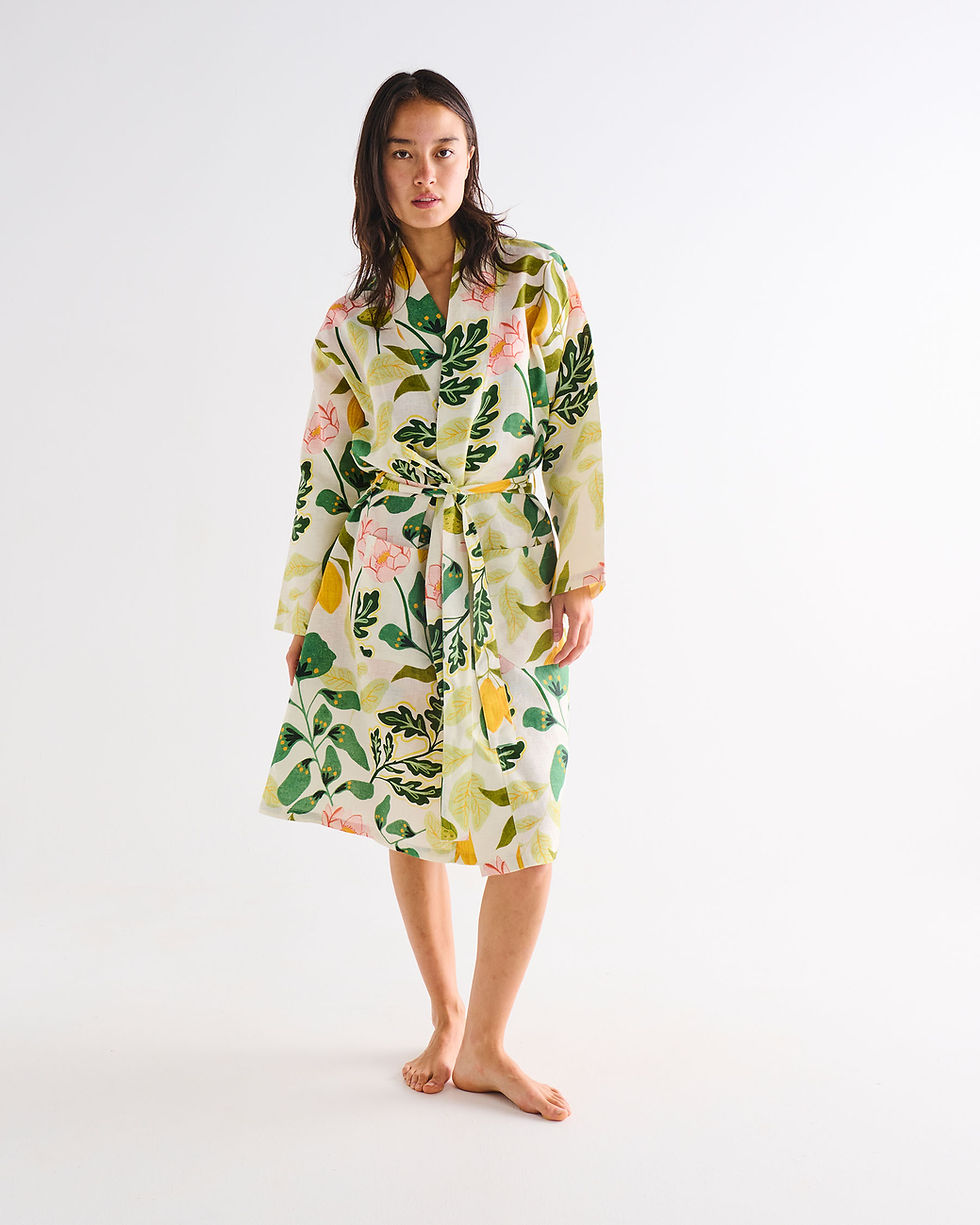 A woman stands barefoot against a plain white studio background wearing a long-sleeve belted robe with a botanical print featuring green leaves, yellow citrus motifs, and pink flowers on a white base.