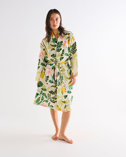A woman stands barefoot against a plain white studio background wearing a long-sleeve belted robe with a botanical print featuring green leaves, yellow citrus motifs, and pink flowers on a white base.