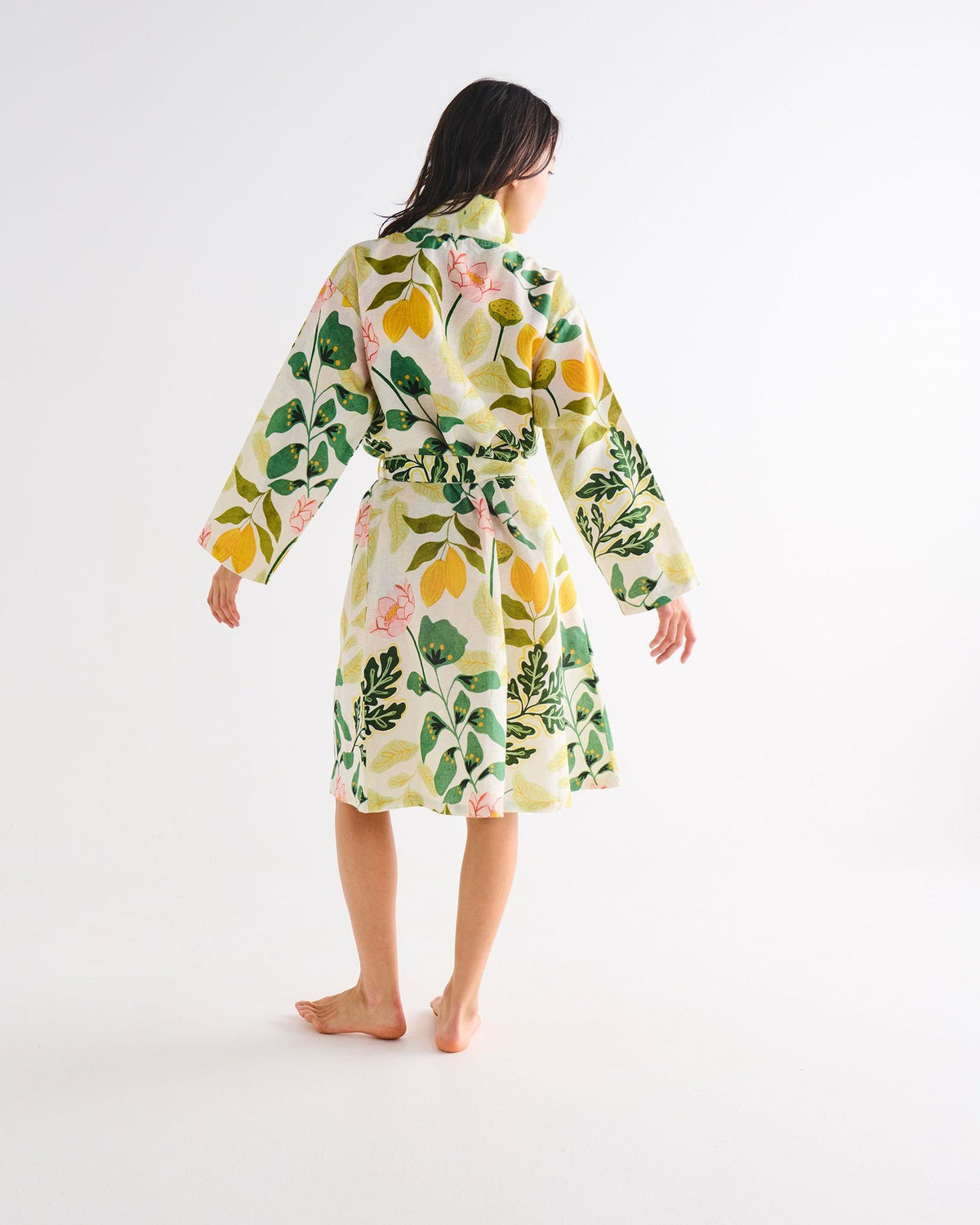 A woman viewed from behind wearing a long-sleeved belted robe with a vibrant green, yellow, and pink botanical print on a white base, standing against a plain white studio background.