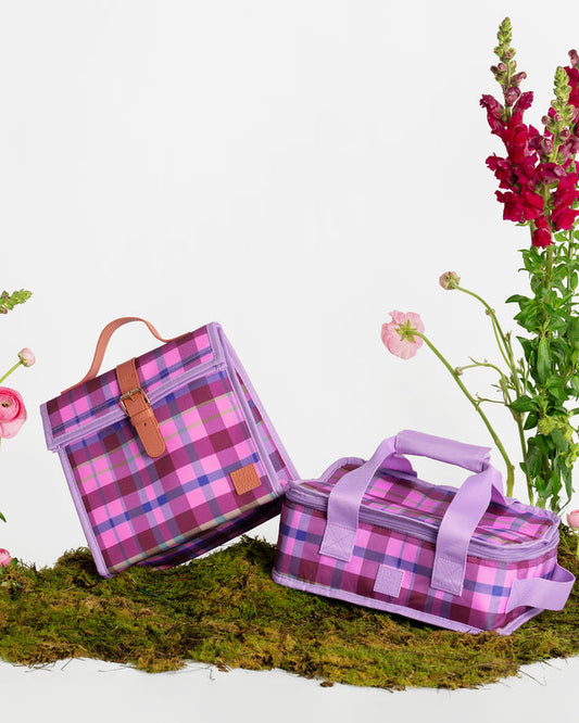 Two purple and pink plaid insulated lunch bags styled on a patch of green moss with flowers against a white background.