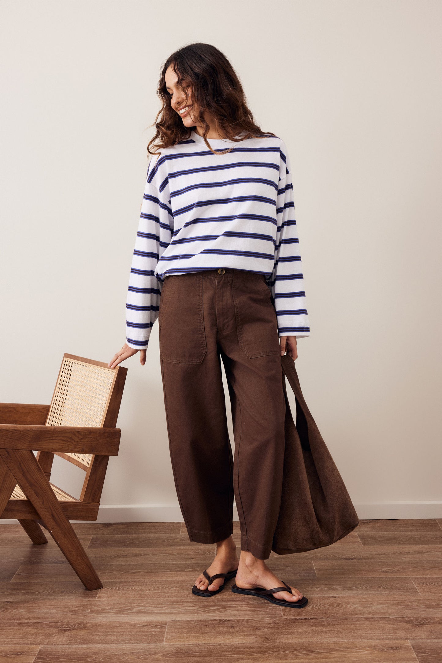 A woman stands in a studio wearing a navy and white horizontally striped long-sleeve top, brown wide-leg cropped pants, and black flip-flops. She is holding a large brown tote bag and standing next to a wooden chair against a light beige wall.