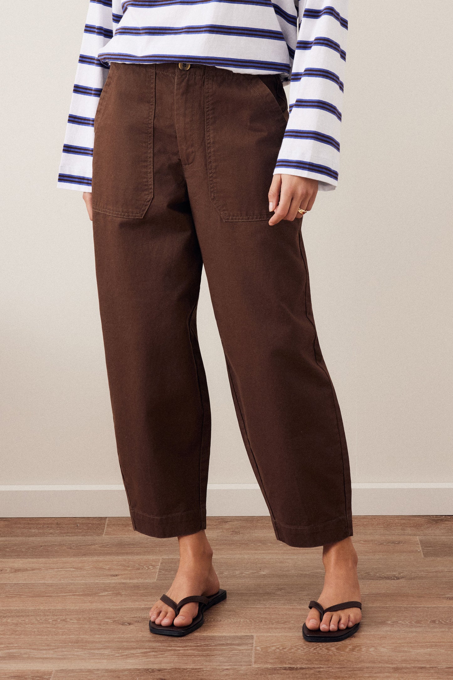 A person wearing dark brown cropped wide-leg pants, a white long-sleeve top with navy blue horizontal stripes, and black flip-flop sandals, standing against a beige wall and light wood floor.