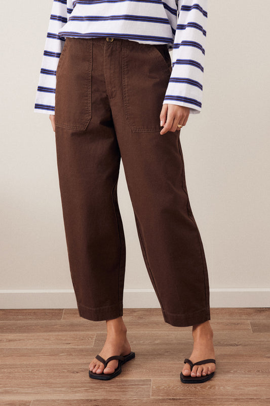 A person wearing dark brown cropped wide-leg pants, a white long-sleeve top with navy blue horizontal stripes, and black flip-flop sandals, standing against a beige wall and light wood floor.