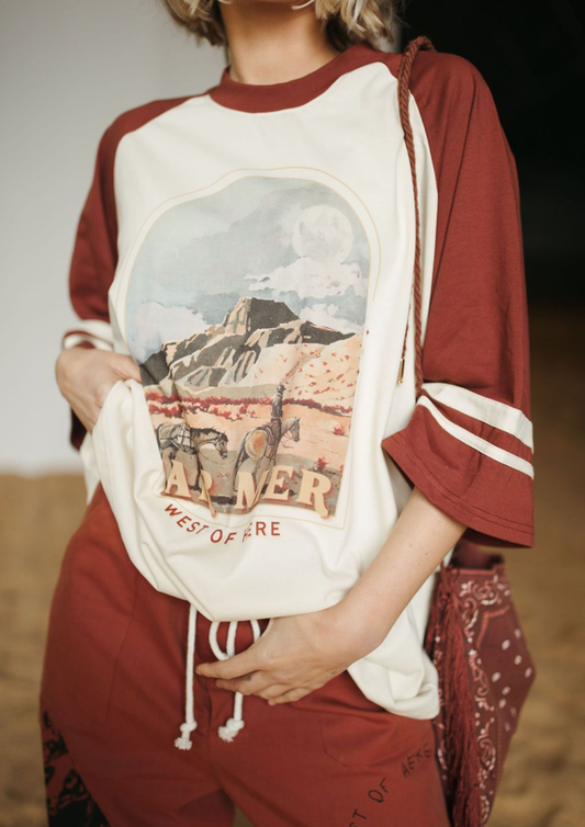 A person wearing a cropped raglan-style T-shirt with a large western desert landscape graphic on the front, paired with matching rust-brown drawstring pants and a bandana bag.