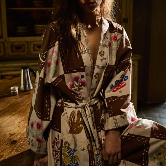A woman wearing a brown and cream patchwork kimono robe with colorful floral prints, tied at the waist, in a rustic indoor setting.